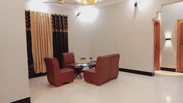 A spacious living room with a high ceiling, a ceiling fan, and a chandelier. There are two red armchairs and a small table with a laptop. The room has a large window with patterned curtains.