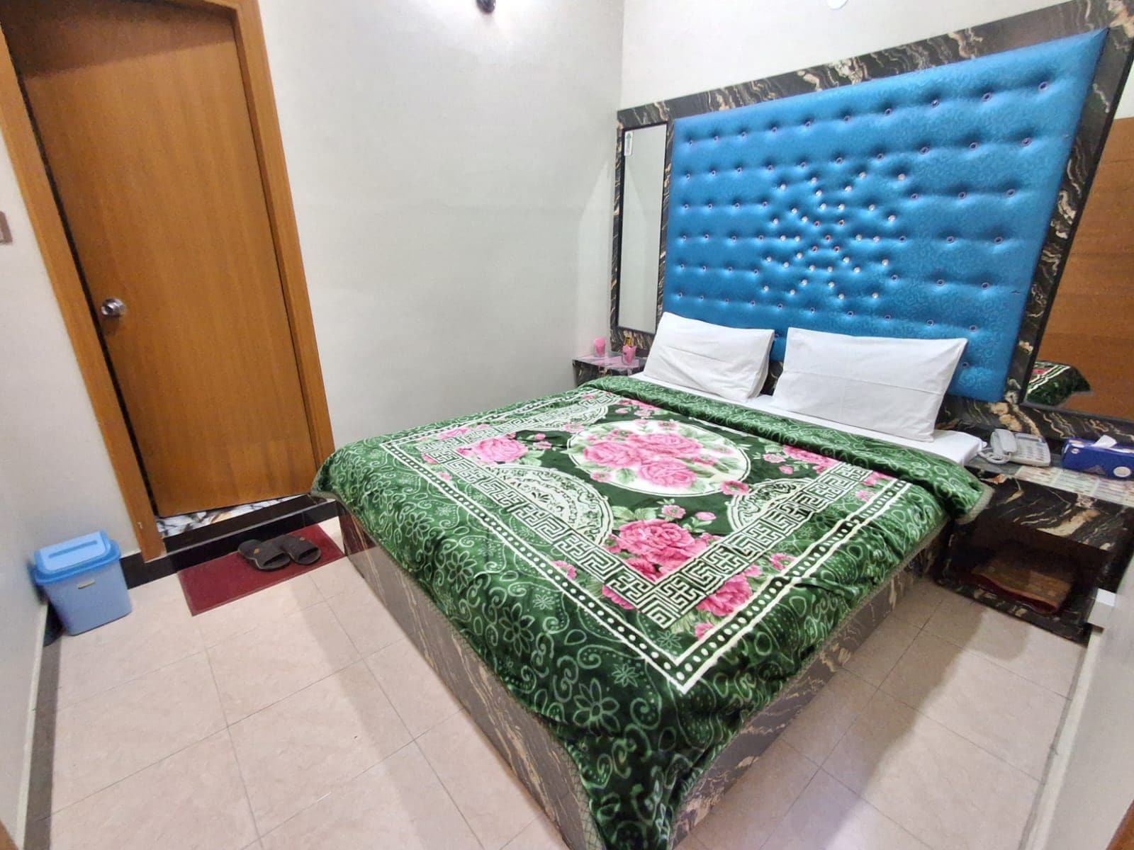 Una habitación con una cama, cabecera azul, mesita de noche, cesto de basura y una puerta.