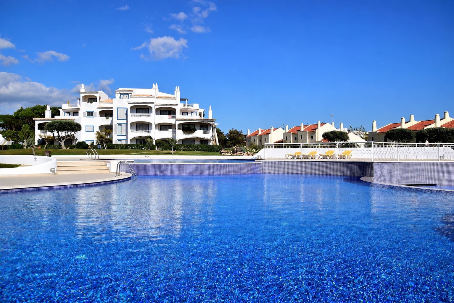 Une piscine à l’eau d’un bleu cristallin, entourée d’une clôture blanche et, au fond, un bâtiment doté de balcons.