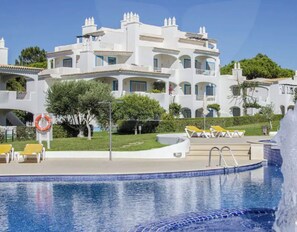 Una piscina con fuente, rodeada de sillas de descanso y un edificio blanco con balcones.