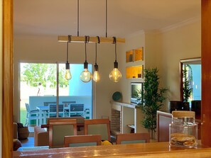 An interior with a wooden ceiling, hanging light fixtures, a dining area with chairs, a table, and a view of the outdoors.