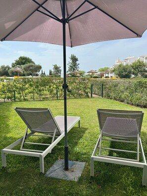 Dos sillones de lounge bajo un paraguas grande en un área verde al aire libre, con un viñedo de fondo.