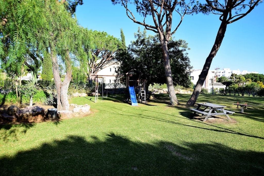 Un espacio al aire libre con césped, que incluye un parque infantil con tobogán y estructura de escalada, rodeado de árboles y una mesa de picnic.