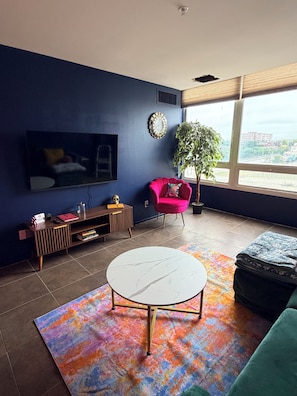 A modern living room with a flat-screen TV, a round coffee table, a vibrant rug, a red chair, and a large window offering a city view. - Midtown Omahas, The Good Life Loft with view of downtown (Omaha)