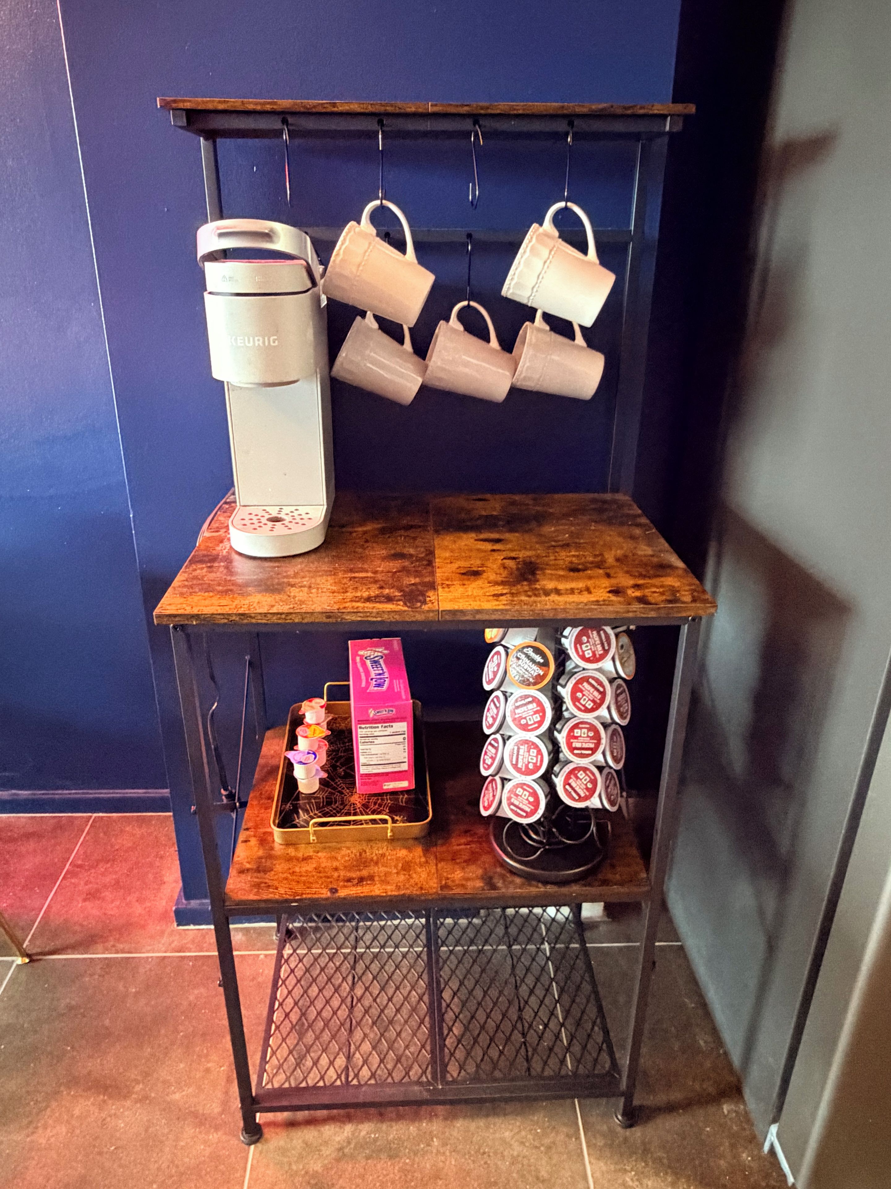 A shelf with a coffee maker, hanging cups, a tray with a box and a small figurine, and a rack of coffee pods.
