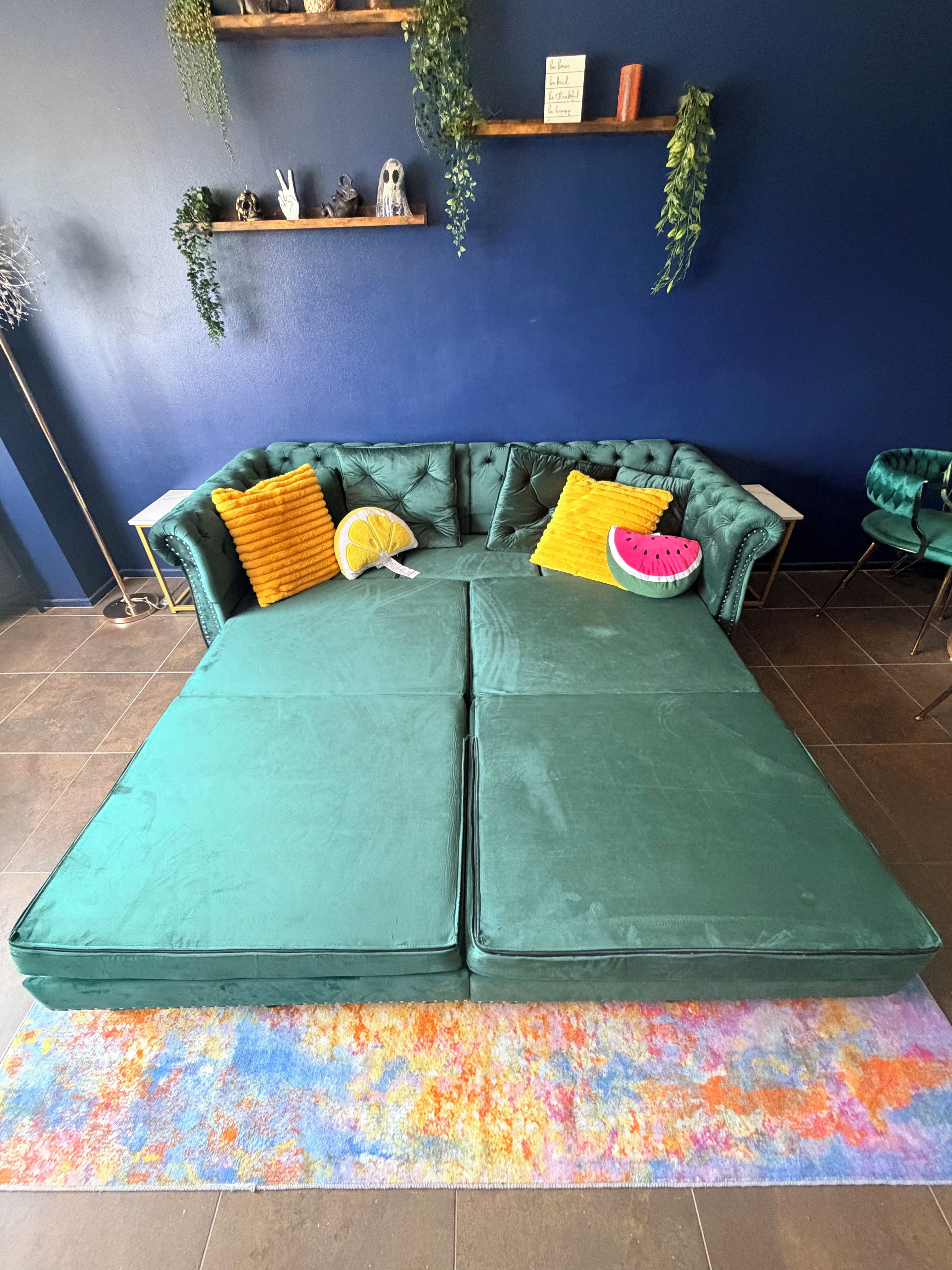 Un sofá seccional verde con dos mesitas auxiliares, una alfombra colorida y plantas decorativas en estanterías.