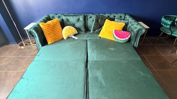 Un sofá seccional verde con dos mesitas auxiliares, una alfombra colorida y plantas decorativas en estanterías.
