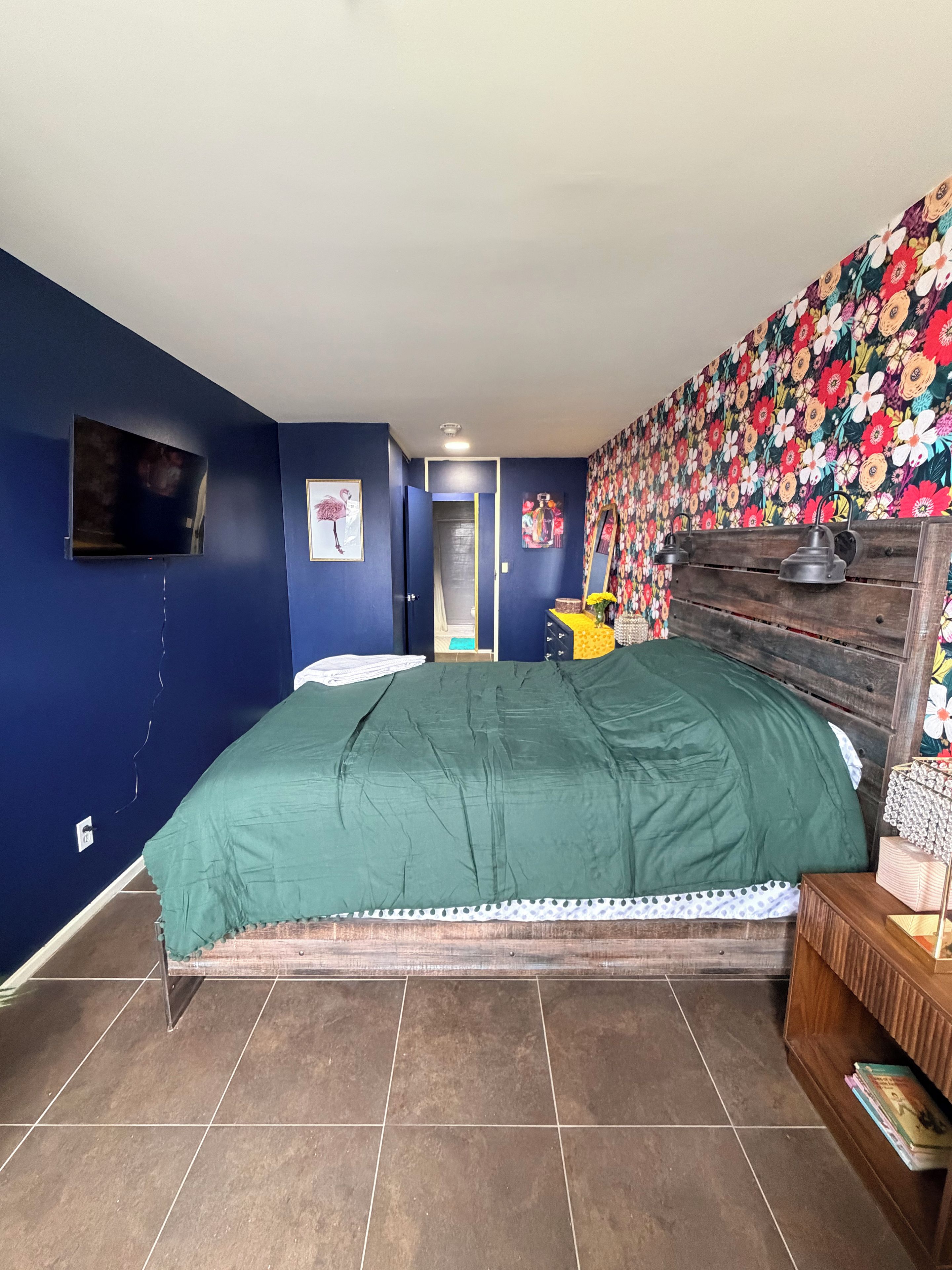Une chambre à coucher avec un lit, un téléviseur fixé au mur, un papier peint à motifs floraux et une tête de lit en bois.