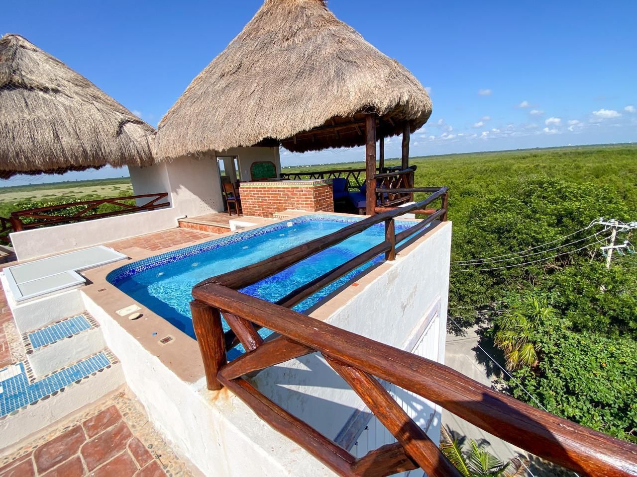 A pool area with a wooden deck, a thatched-roof structure, and a view of a green landscape.