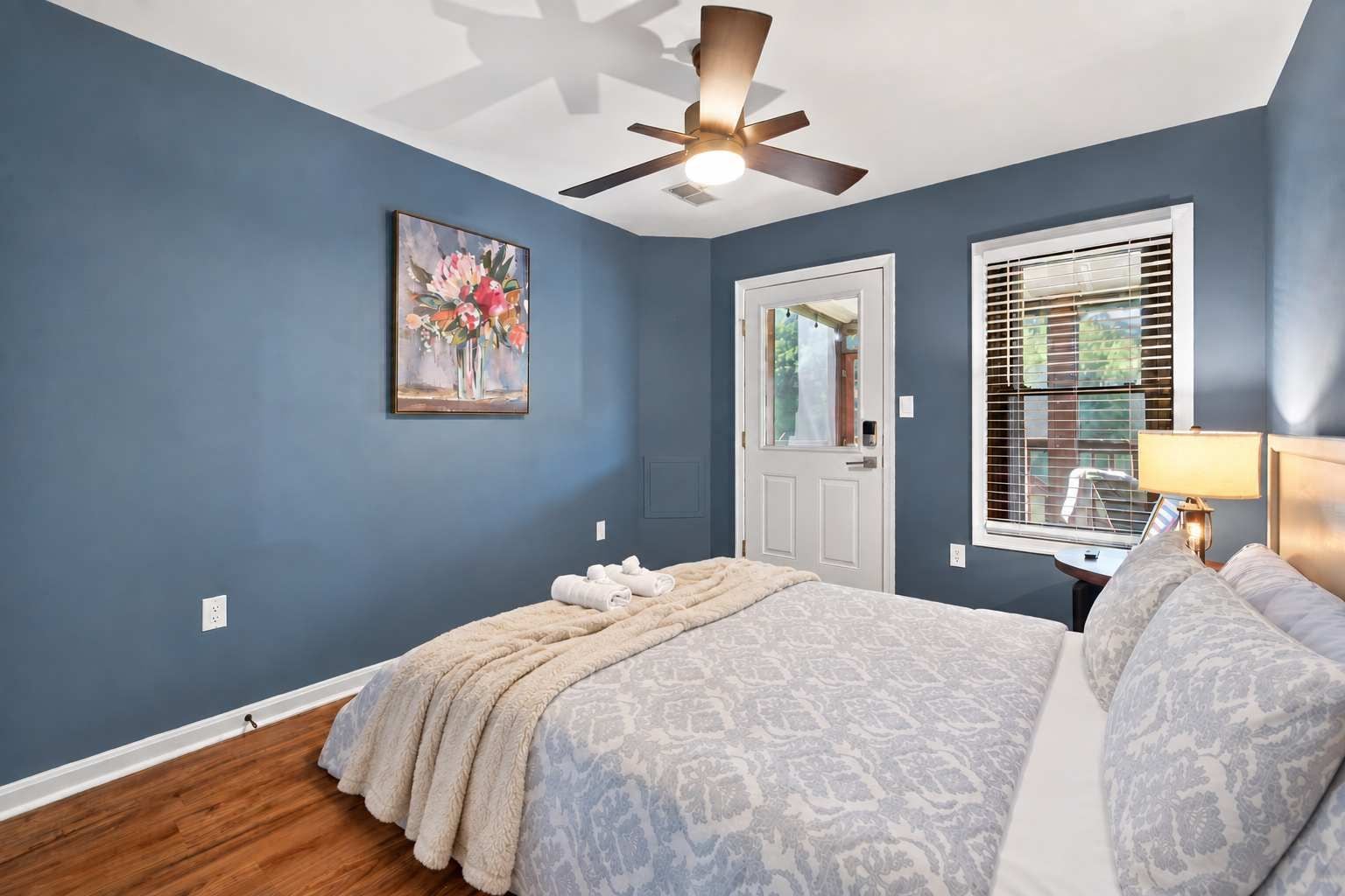 A bedroom with a bed, a ceiling fan, a door, a window with blinds, and a painting on the wall.