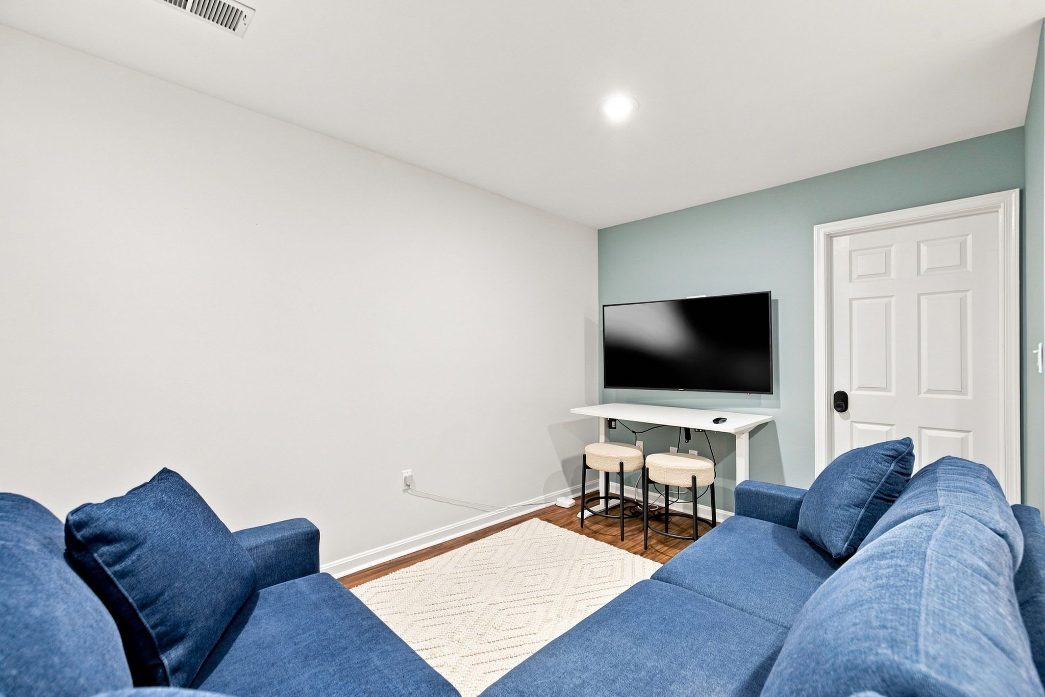 A living room with a sofa set, a flat-screen TV, a small table with two chairs, and a white door.