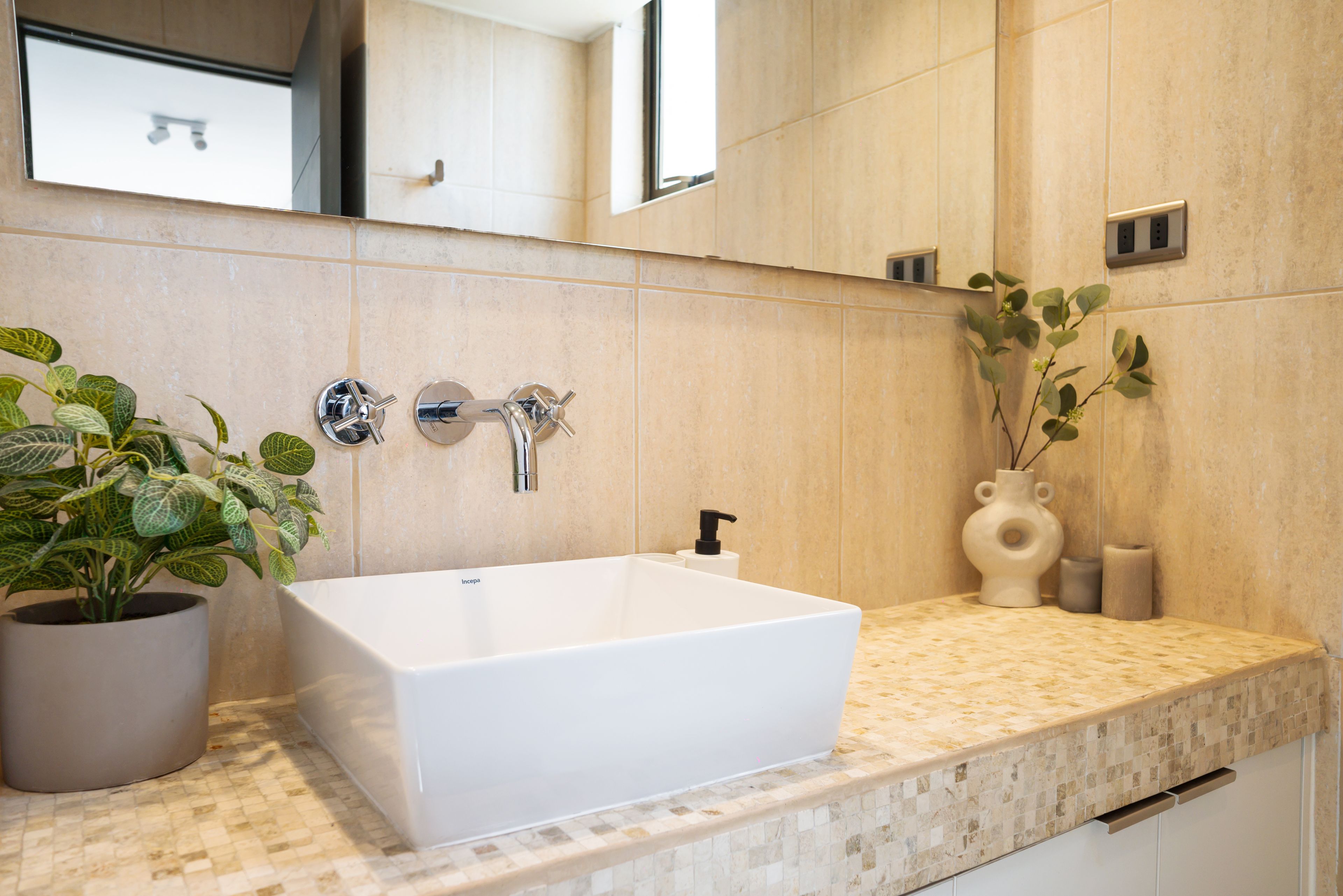 Un bagno con un lavandino bianco, uno specchio, una pianta in vaso e un vaso con fiori.