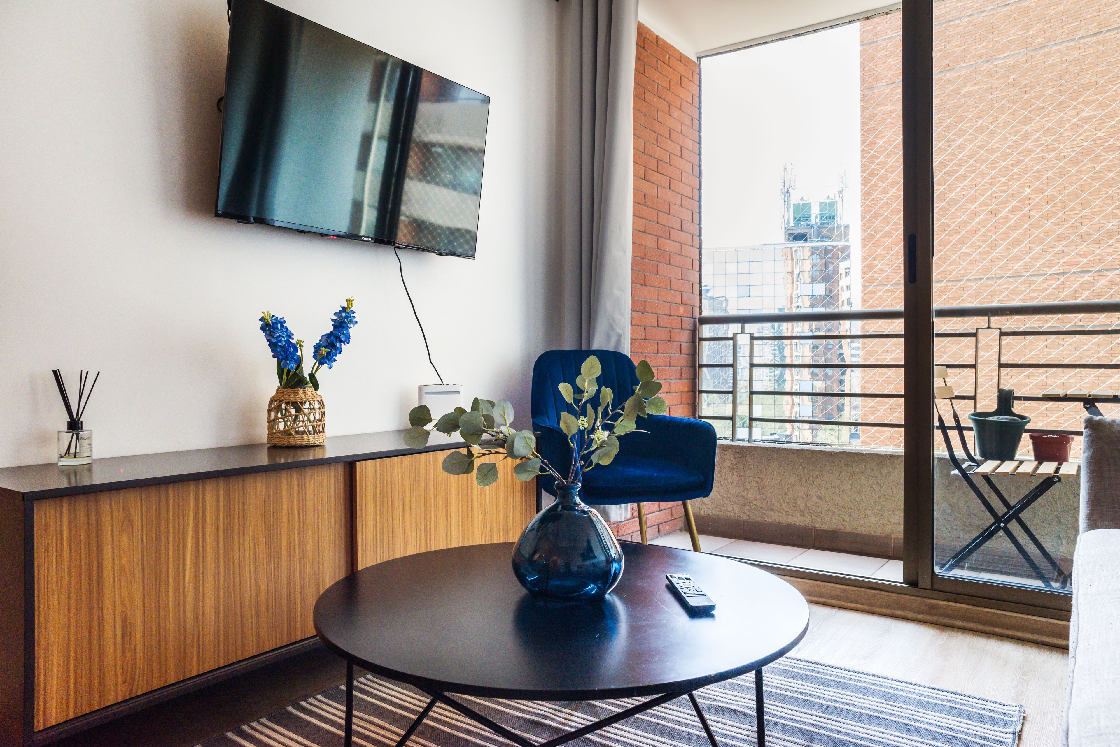 Habitación de hotel moderna con un televisor de pantalla plana montado en la pared, una mesa redonda con un jarrón de flores, una silla azul y un balcón con vistas a la ciudad.