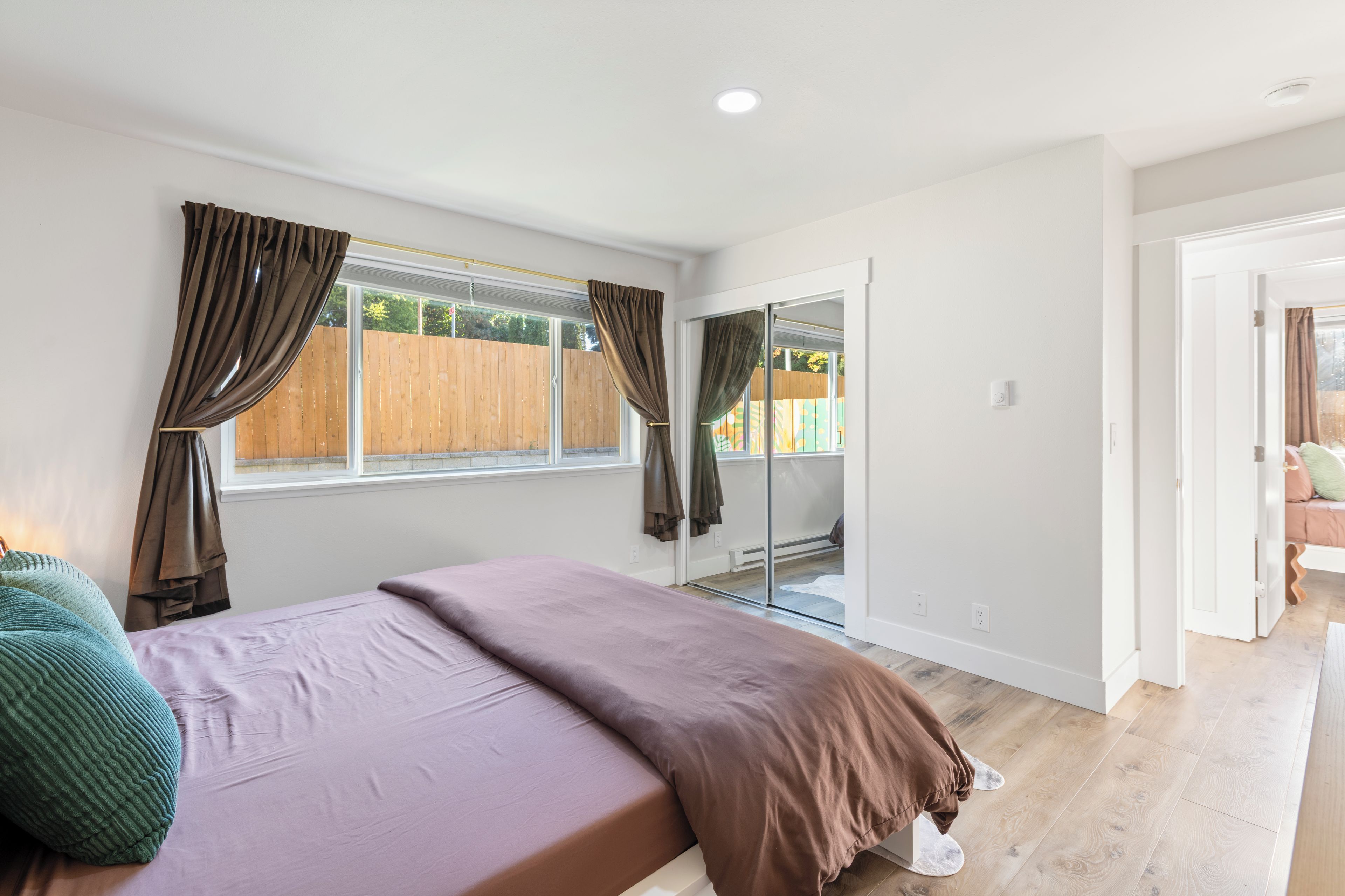 A bedroom with a bed, a window with curtains, a sliding glass door, and a view of a wooden fence outside.
