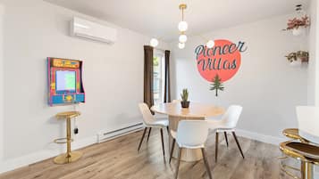 Een moderne kamer met een eettafel, stoelen, een wandairconditioner, een retro speelautomaat en een muursticker.