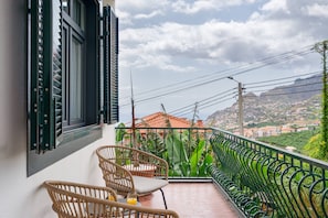 A balcony with wicker chairs, a table, and a view of a town with houses and mountains. - Casa dos Amigos by Galmi (Câmara de Lobos)
