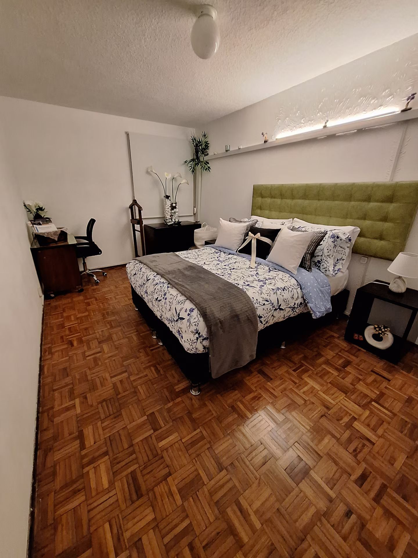 Un dormitorio con suelo de parqué de madera, una cama con ropa de cama floral, un cabecero tapizado en verde, una mesita de noche con lámpara y una estantería con objetos decorativos.