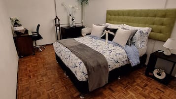Un dormitorio con suelo de parqué de madera, una cama con ropa de cama floral, un cabecero tapizado en verde, una mesita de noche con lámpara y una estantería con objetos decorativos.