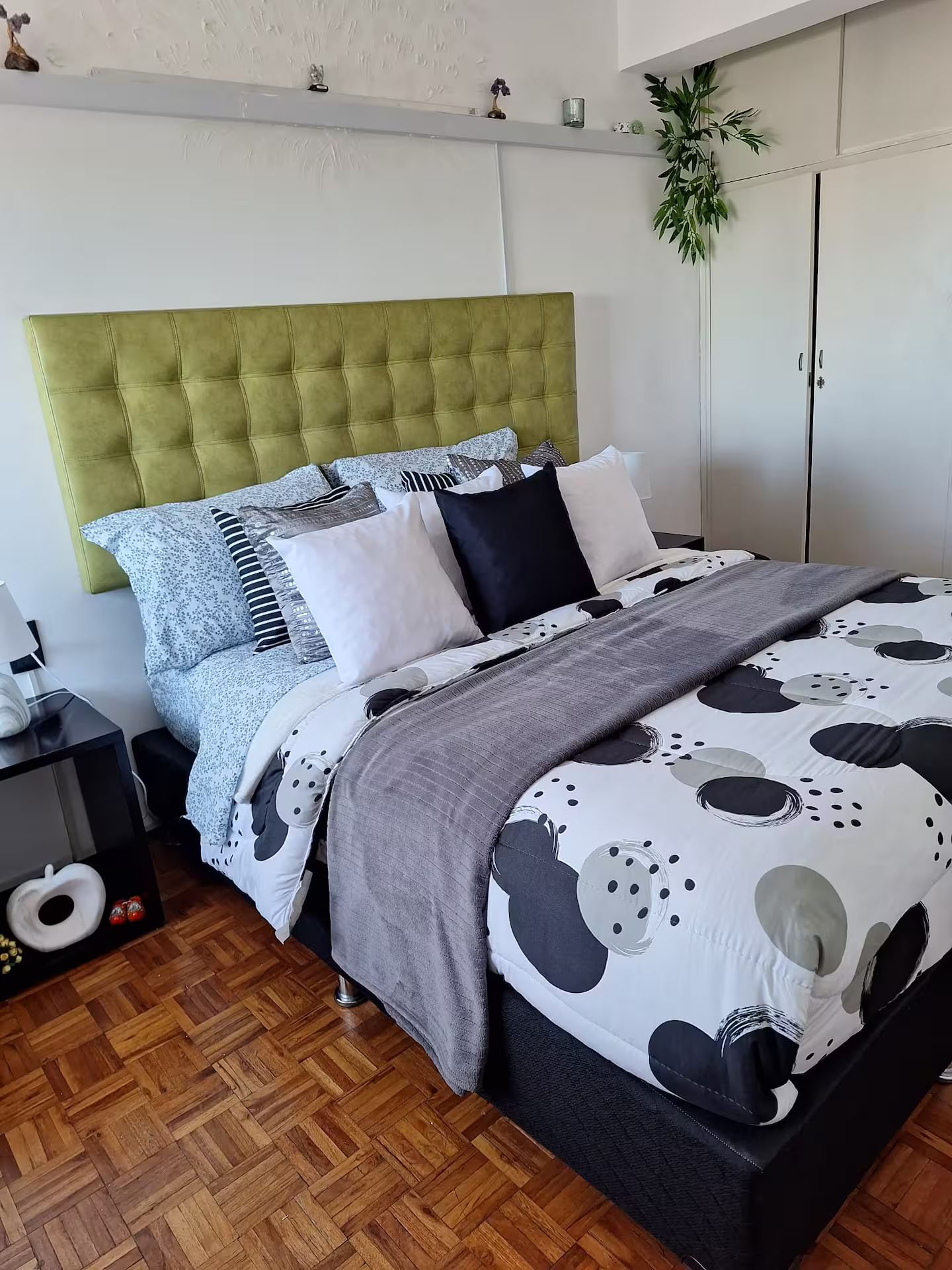 A modern bedroom with a tufted green headboard, a patterned bedspread, and a wooden parquet floor.