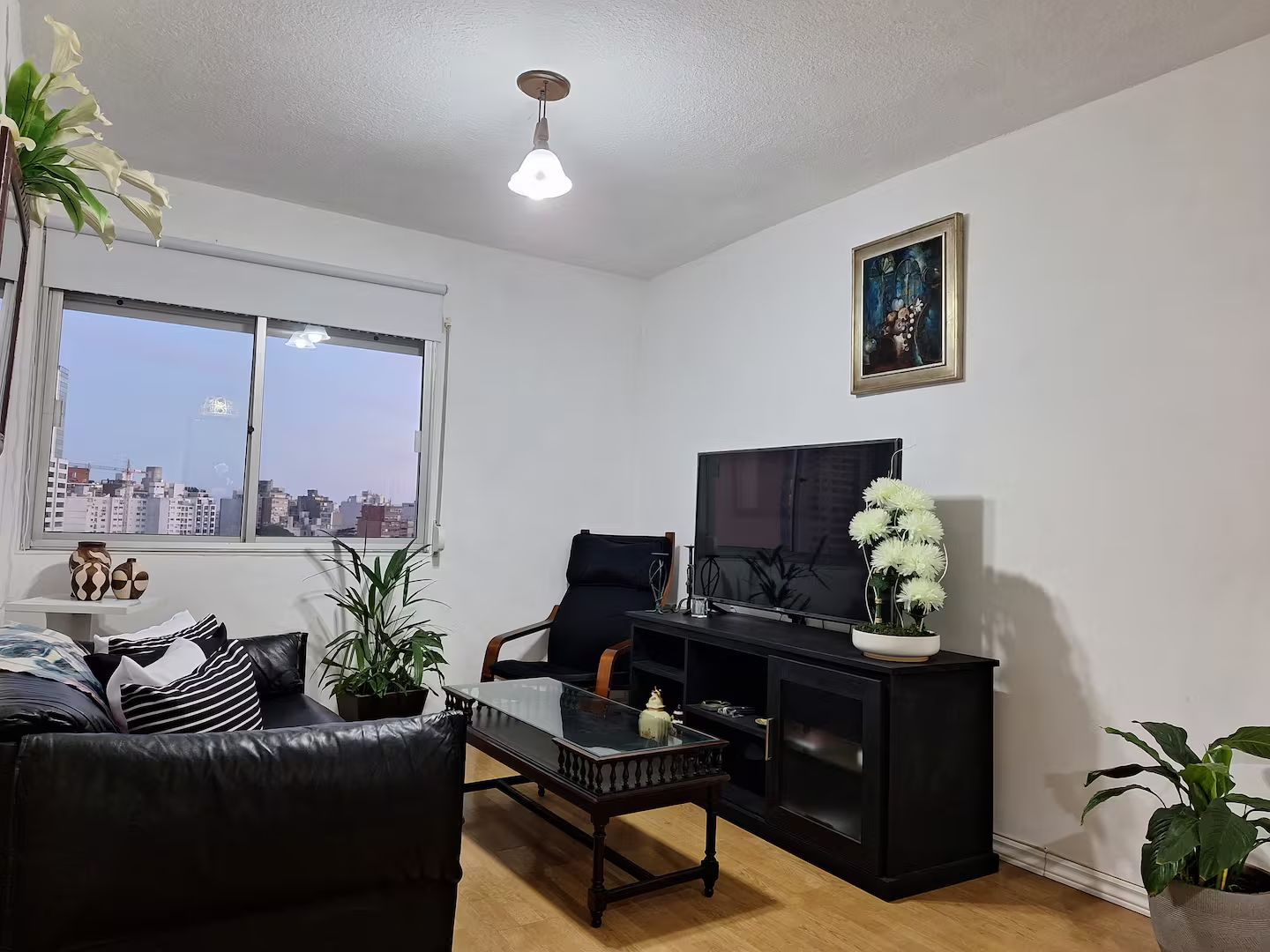 A living room with a black leather sofa, a glass coffee table, a black entertainment unit with a TV, and a window with a city view.