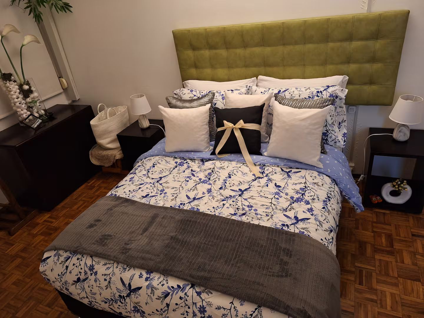 Una cama con colcha estampada con motivos florales, cabecera tapizada de verde y varias almohadas.