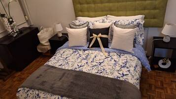 Una cama con colcha estampada con motivos florales, cabecera tapizada de verde y varias almohadas.