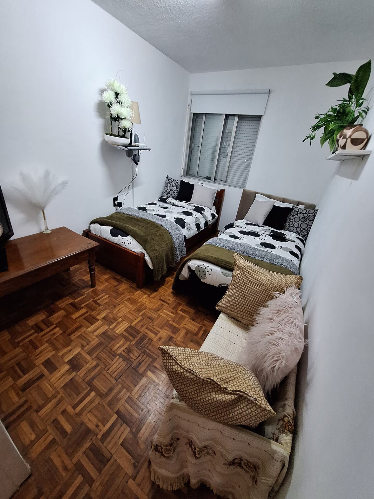 A compact bedroom with two single beds, a wooden floor, a small table, and a wall-mounted shelf with a plant.