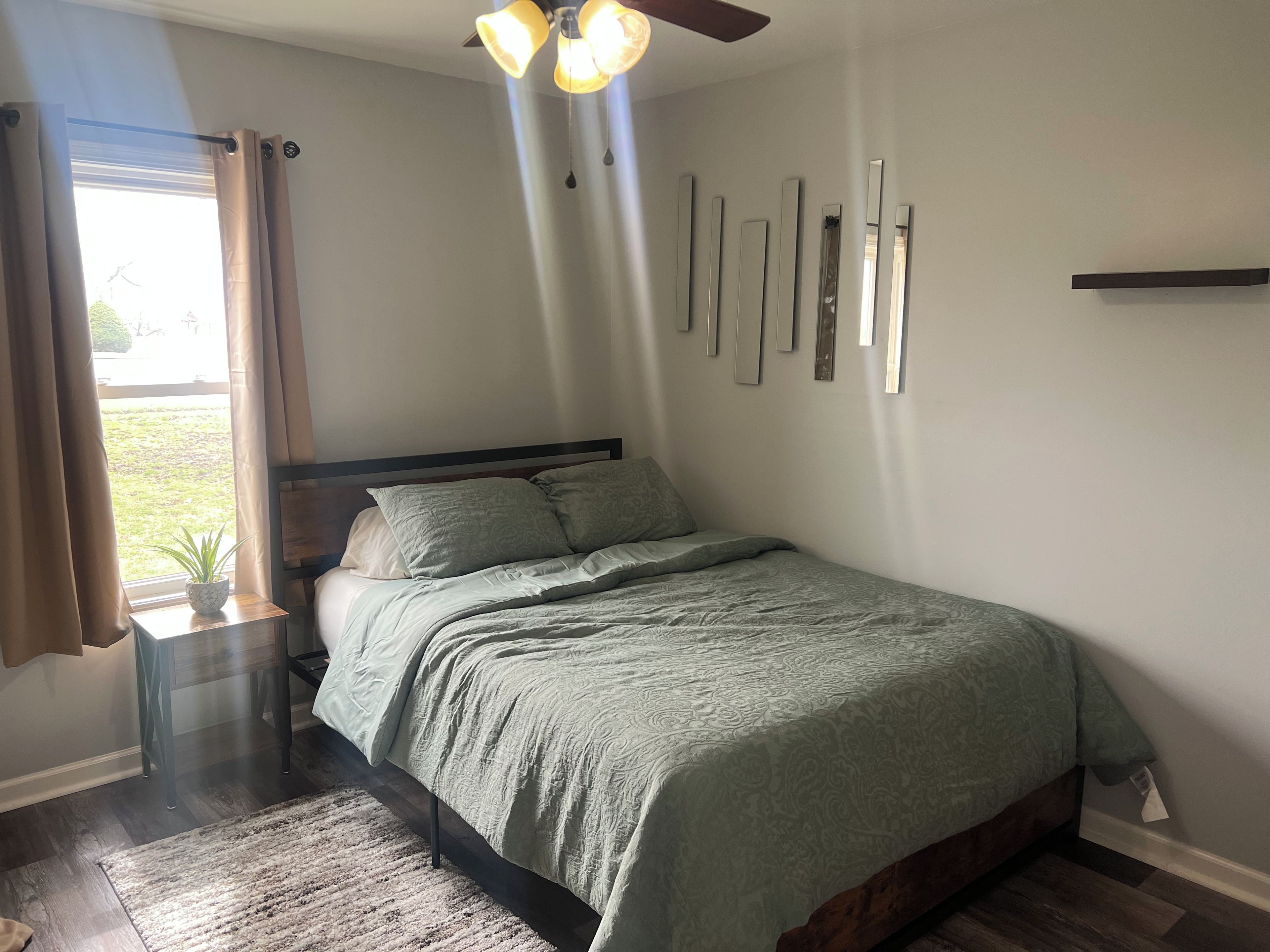 Un dormitorio con cama, ventilador de techo, ventana con cortinas y espejo montado en la pared.