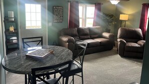 A living room with a brown leather couch, a brown leather armchair, a glass-top coffee table, a dark wooden side table, and a wall-mounted sign with inspirational text.