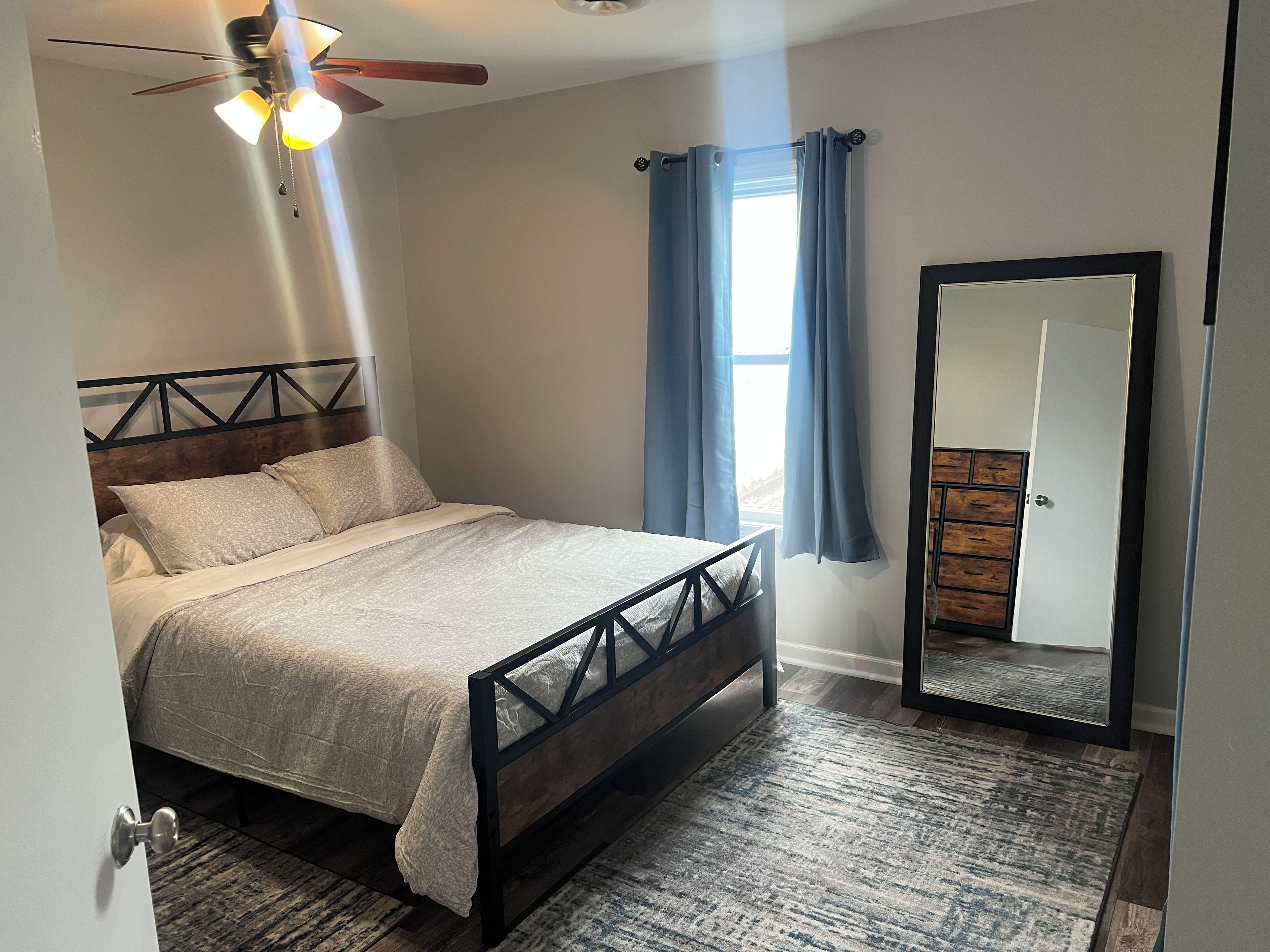Un dormitorio con cama, ventilador de techo, ventana con cortinas, espejo y armario.