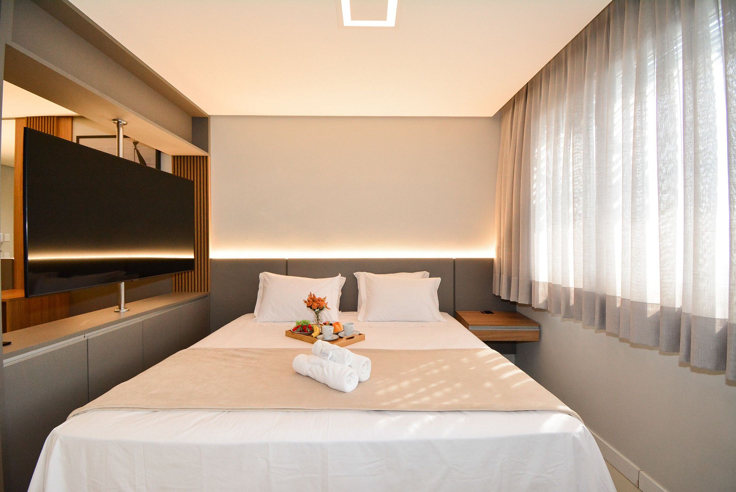 Habitación de hotel con una cama grande, un televisor de pantalla plana montado en la pared, una mesita de noche y un ventanal con cortinas traslúcidas.