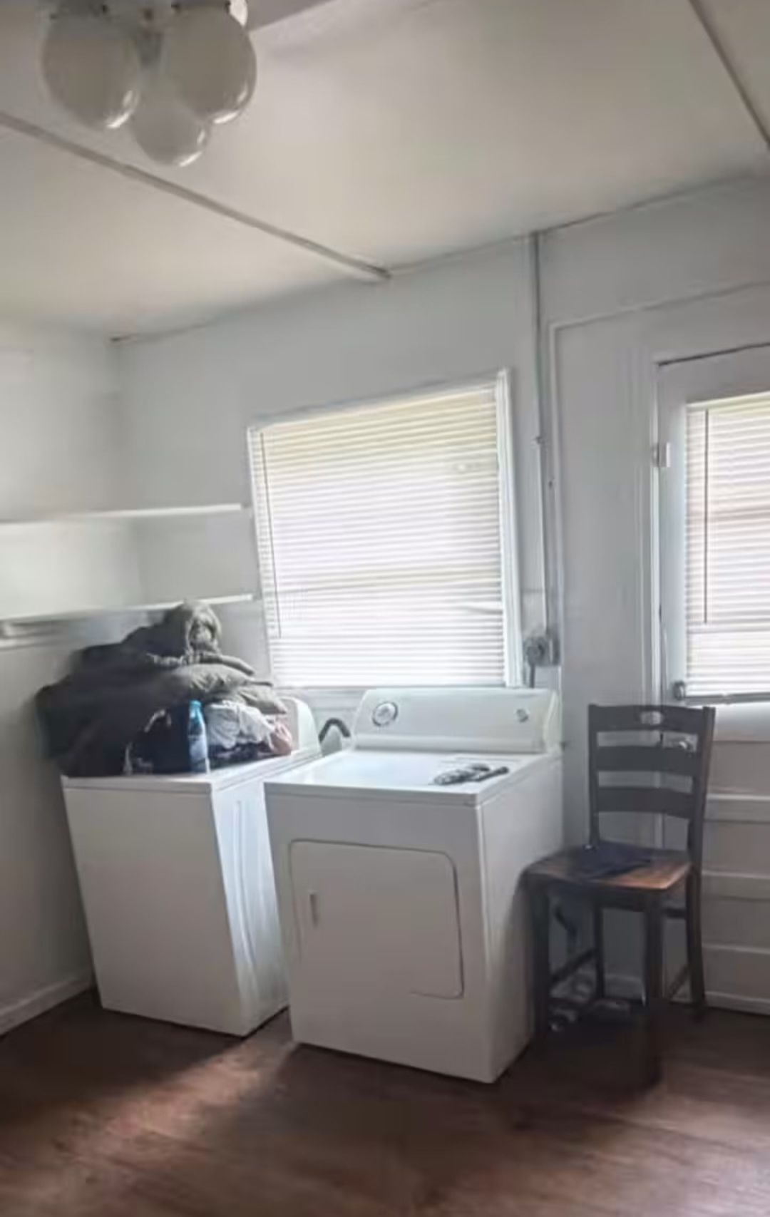 A laundry room with a washing machine and dryer, a chair, and a window with blinds.