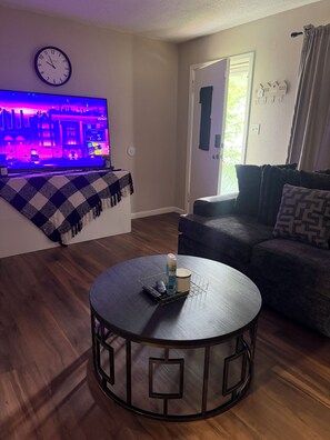 A living room with a round table, a sofa, a television, and a door with a number. - Cozy modern ranch retreat 2BR & 1BA (St. Louis)