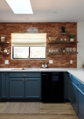 A kitchen with a brick backsplash, a window with a roller blind, and built-in shelves with decorative items. - Red Rock Views, Rooftop Patio, Hot Tub, Updated (Sedona)