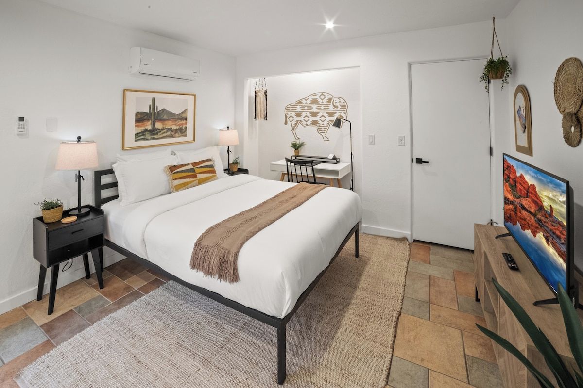 A bedroom with a bed, bedside tables, a desk, a chair, a TV, and a door.