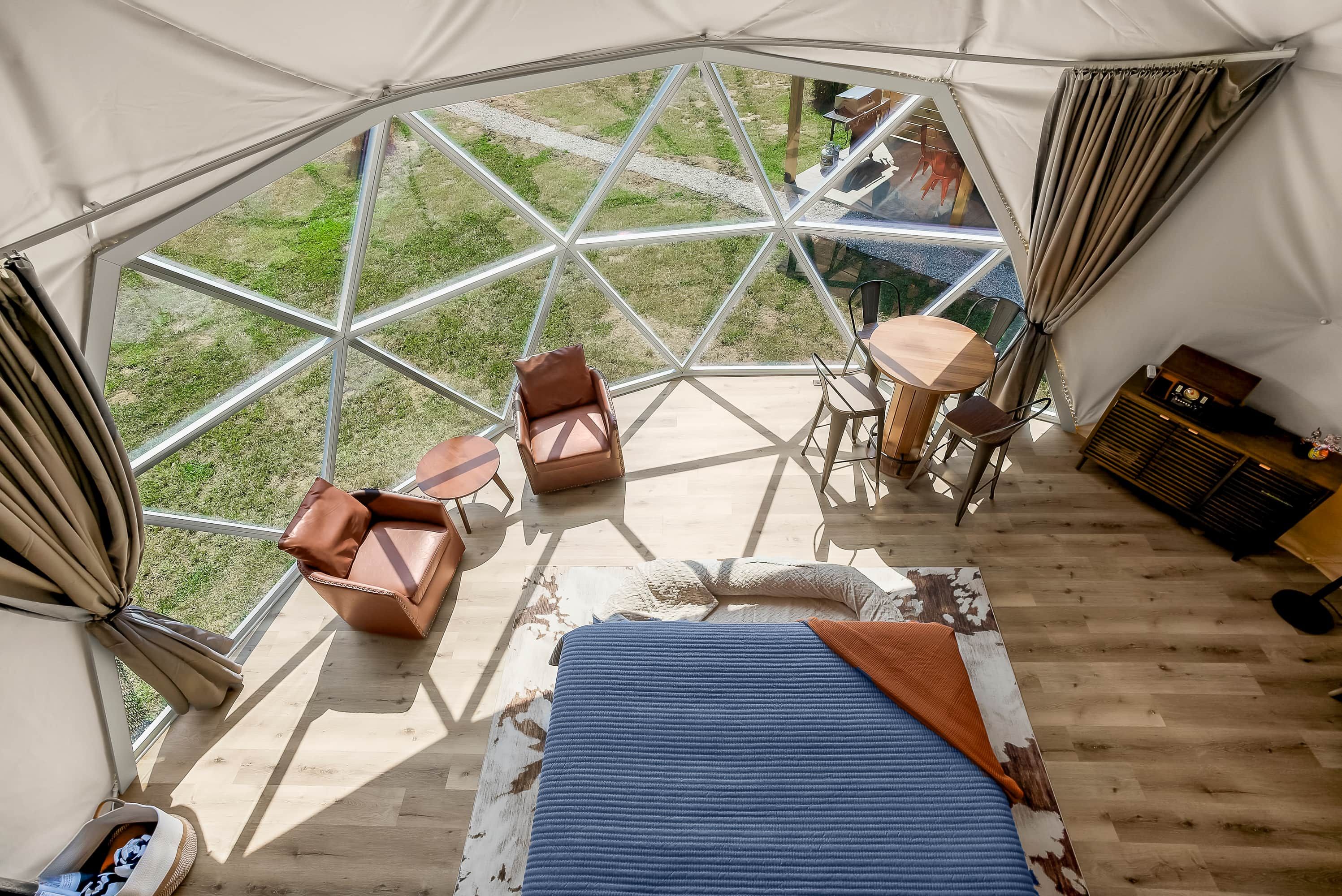 Una carpa con techo de cristal geométrico, amueblada con sillones de cuero, una mesita y una cama con cubrecama azul.