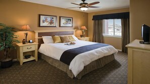 Une chambre d’hôtel avec un grand lit, deux tables de chevet avec des lampes, un ventilateur de plafond, une télévision et une fenêtre avec des rideaux.