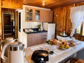 A kitchen with wooden cabinets, a refrigerator, a coffee machine, and a dining table with fruits and pastries.