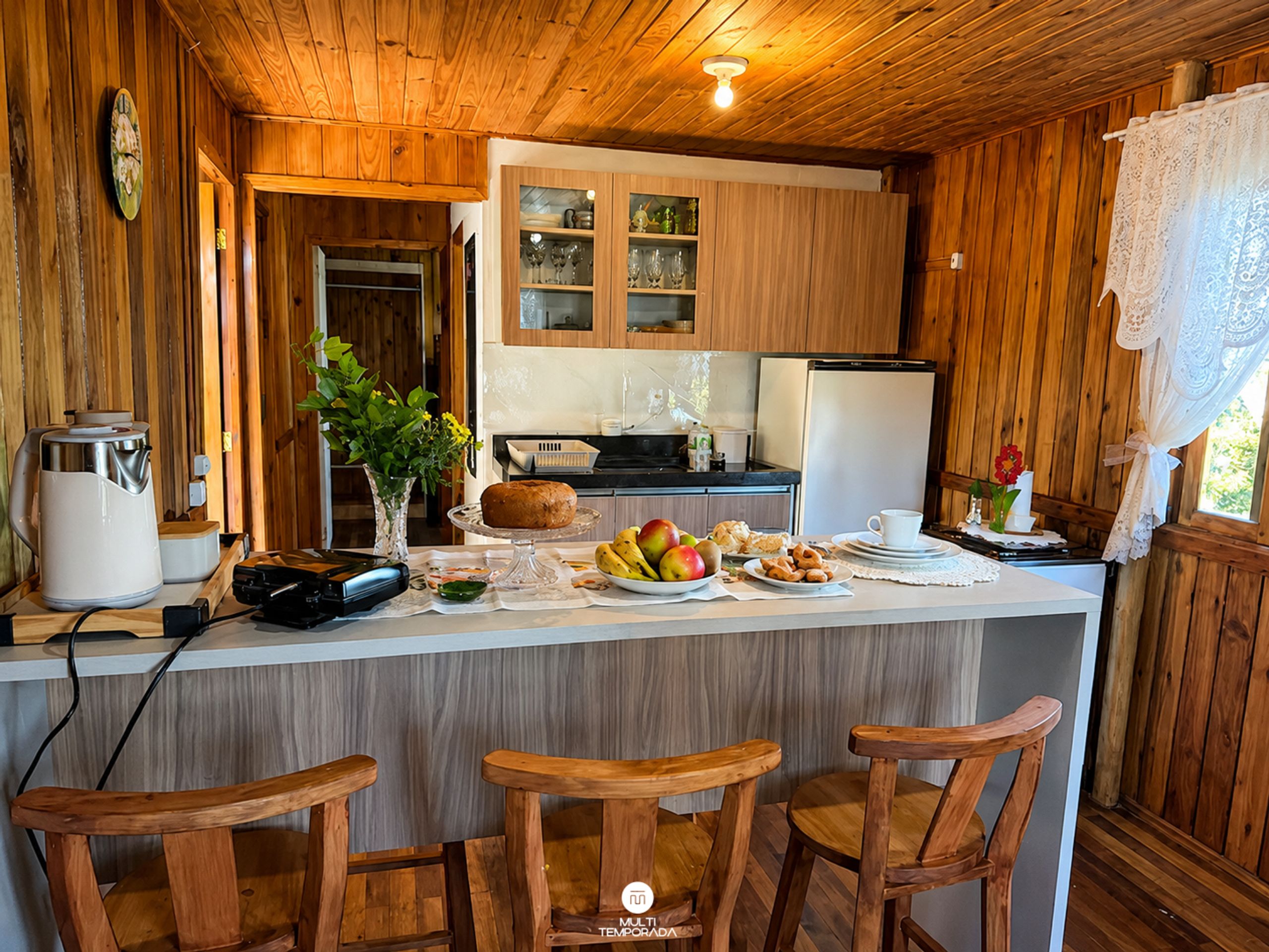 A wooden interior with a kitchen area, including a refrigerator, a coffee maker, and a fruit bowl on the counter.
