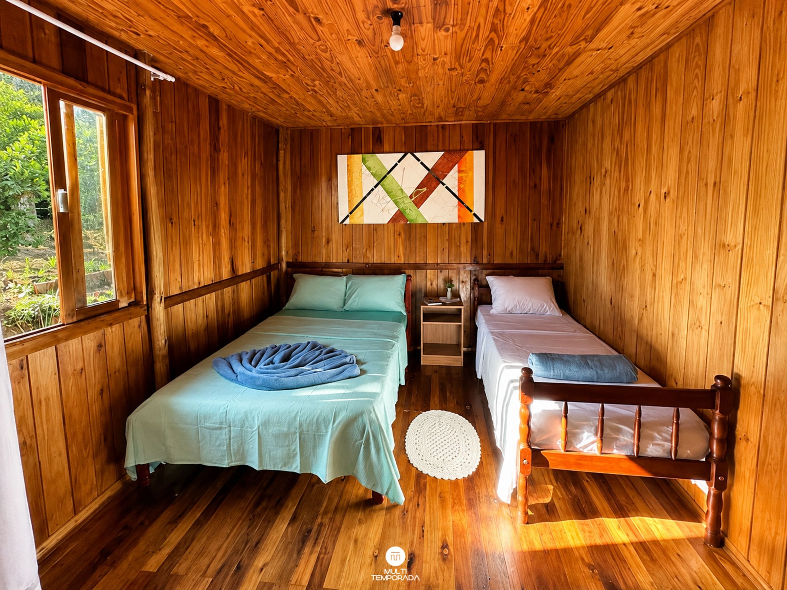 A wooden cabin bedroom with two beds, a nightstand, a window, and a wall-mounted light fixture.