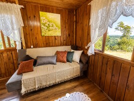Interior de cabana de madeira com sofá, almofadas, manta, cortinas de renda e pintura de flores.