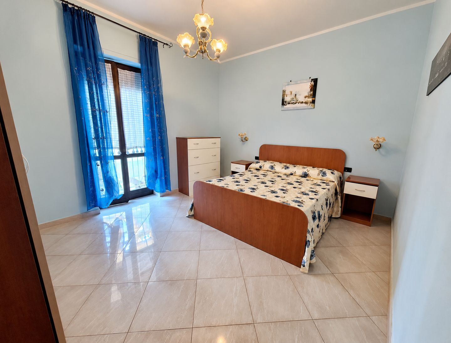 Une chambre à coucher comprenant un lit, une commode, une table de chevet, un lustre et une fenêtre avec des rideaux.