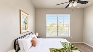 Un dormitorio con cama, lámparas de noche, ventilador de techo, ventana con persianas y un cuadro en la pared.