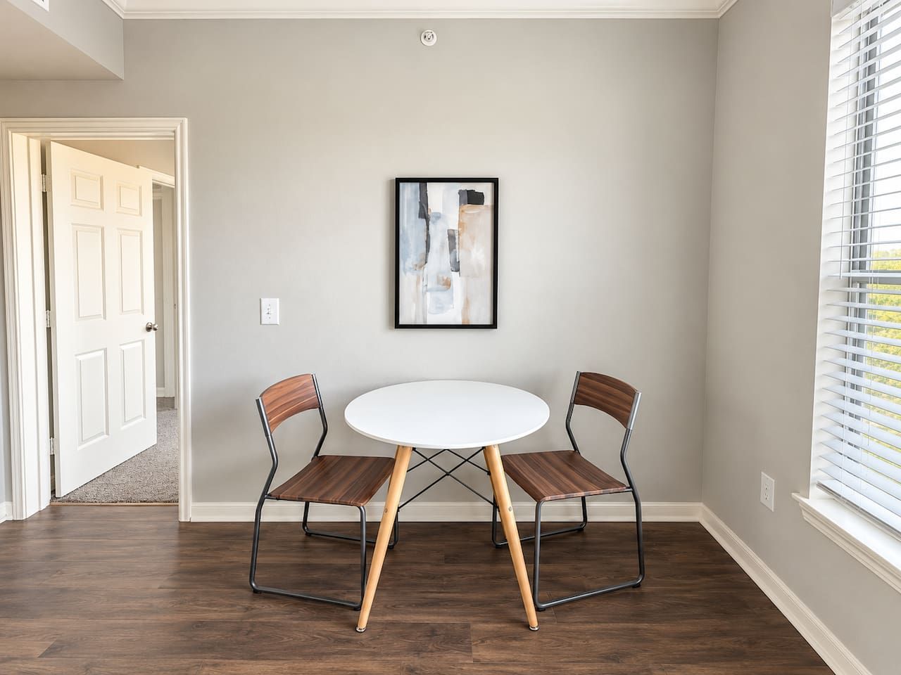 A dining area with a round table and two chairs, a framed abstract painting on the wall, and a window with blinds.