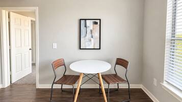 A dining area with a round table and two chairs, a framed abstract painting on the wall, and a window with blinds.