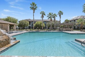 A clear swimming pool with a waterfall feature, surrounded by palm trees and a covered seating area. - Sleepover | Serene 1BD/1BA + Gym - Corpus Christi (Corpus Christi)