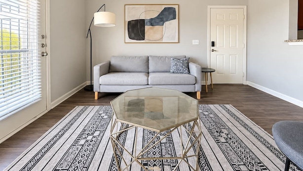 Una sala de estar con una mesa de centro de vidrio, un sofá, una lámpara de pie y una alfombra estampada.