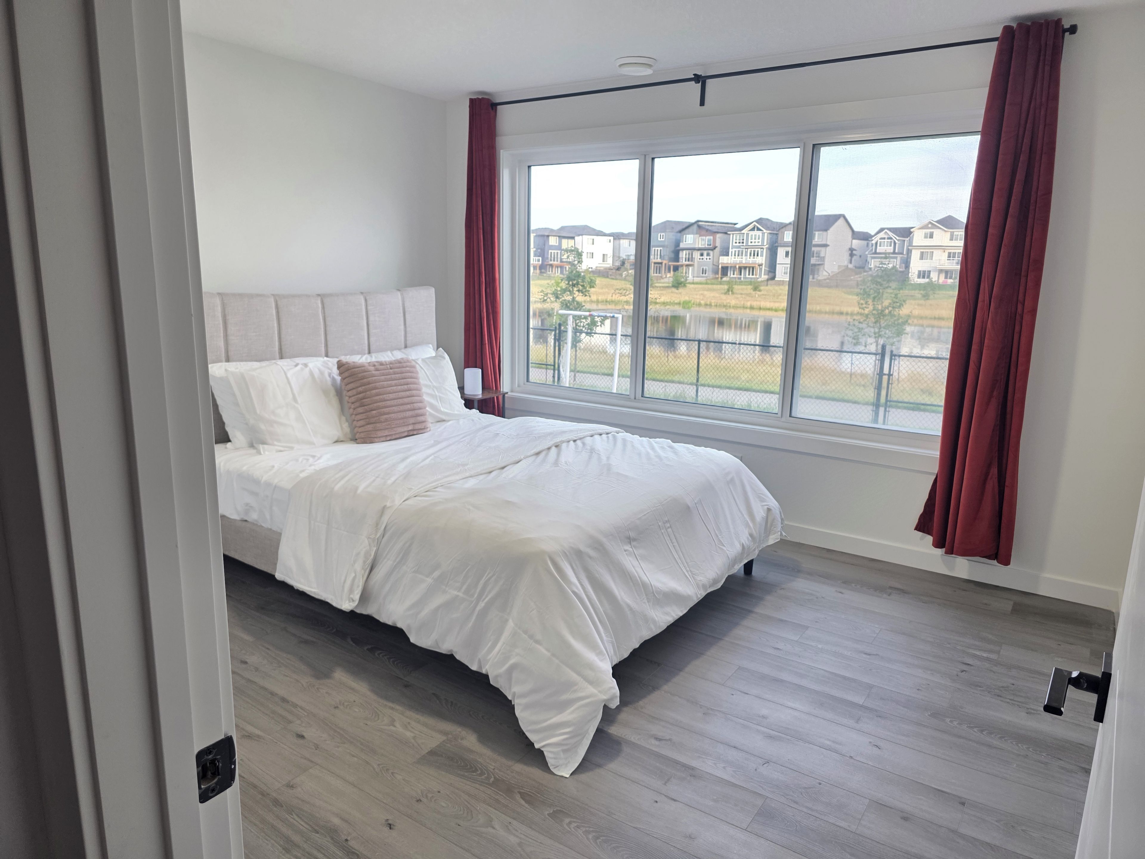 A bedroom with a large window, a bed, and a view of a residential area.