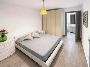 Un dormitorio con cama, mesitas de noche, armario, lámpara colgante y baño visible a través de una puerta abierta.