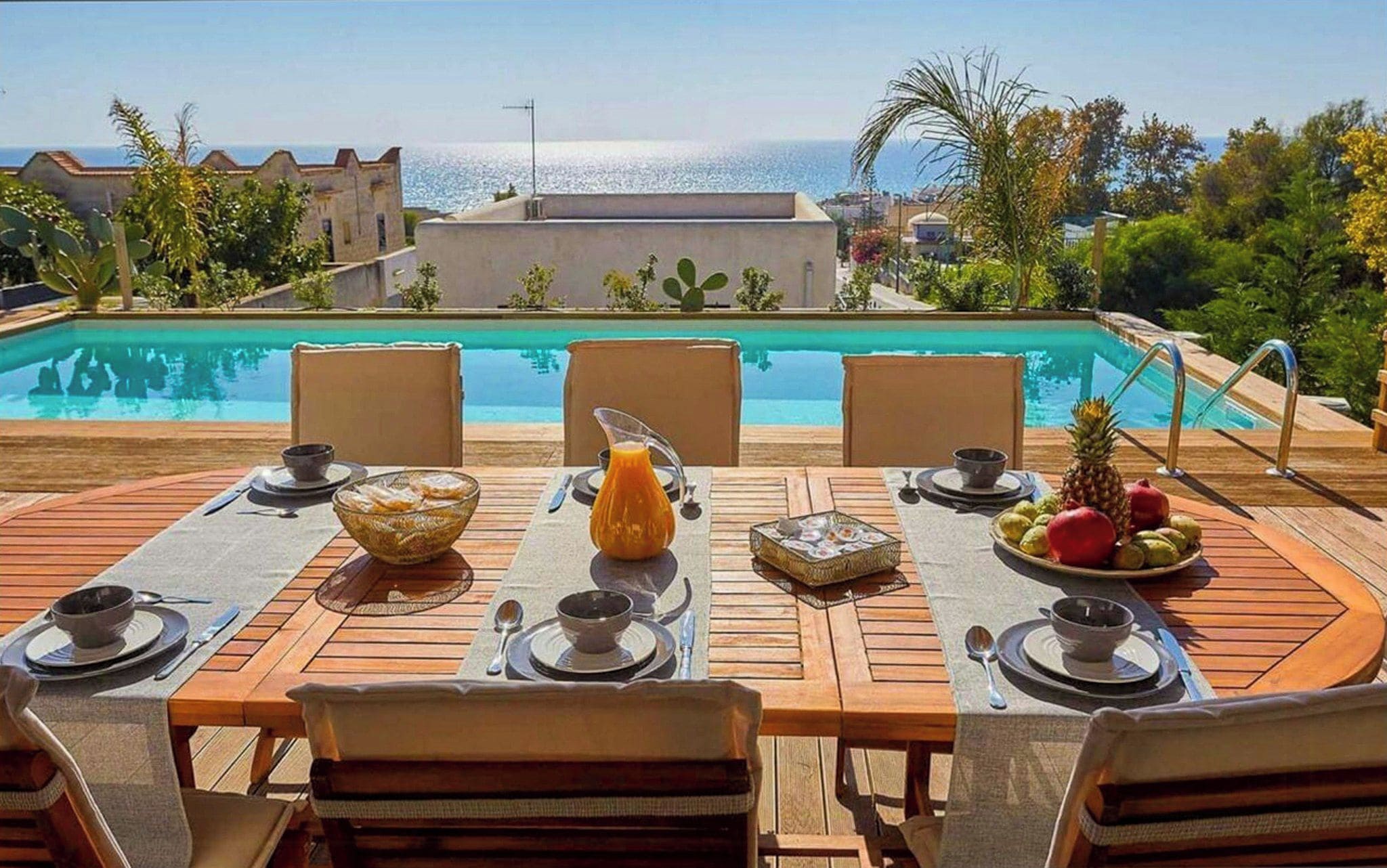 Un área de comedor al aire libre con vista a la piscina, mesas y sillas de madera, y una exhibición de frutas tropicales.